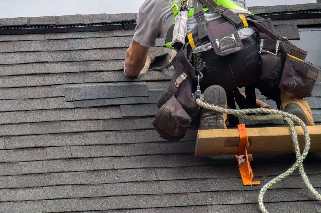 Homeowner repairing roof shingles with safety equipment on a sunny day